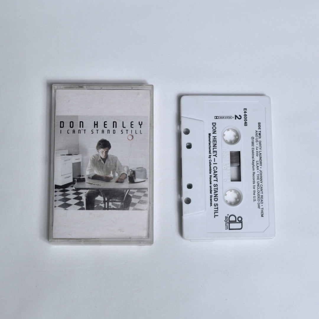 Don Henley – I Can't Stand Still cassette tape, cover photo of a man seated at a kitchen table with a toaster and checkered floor, shown alongside the white cassette (Side 2)