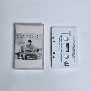 Don Henley – I Can't Stand Still cassette tape, cover photo of a man seated at a kitchen table with a toaster and checkered floor, shown alongside the white cassette (Side 2)