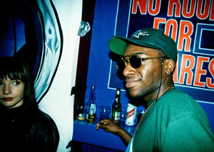 Man wearing sunglasses and a green cap in a nightclub with a blue wall and text.