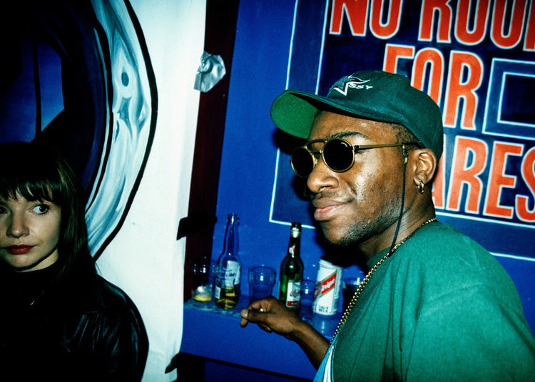 Man wearing sunglasses and a green cap in a nightclub with a blue wall and text.