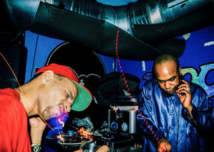 Two men in a club setting with DJ equipment and colorful lights.