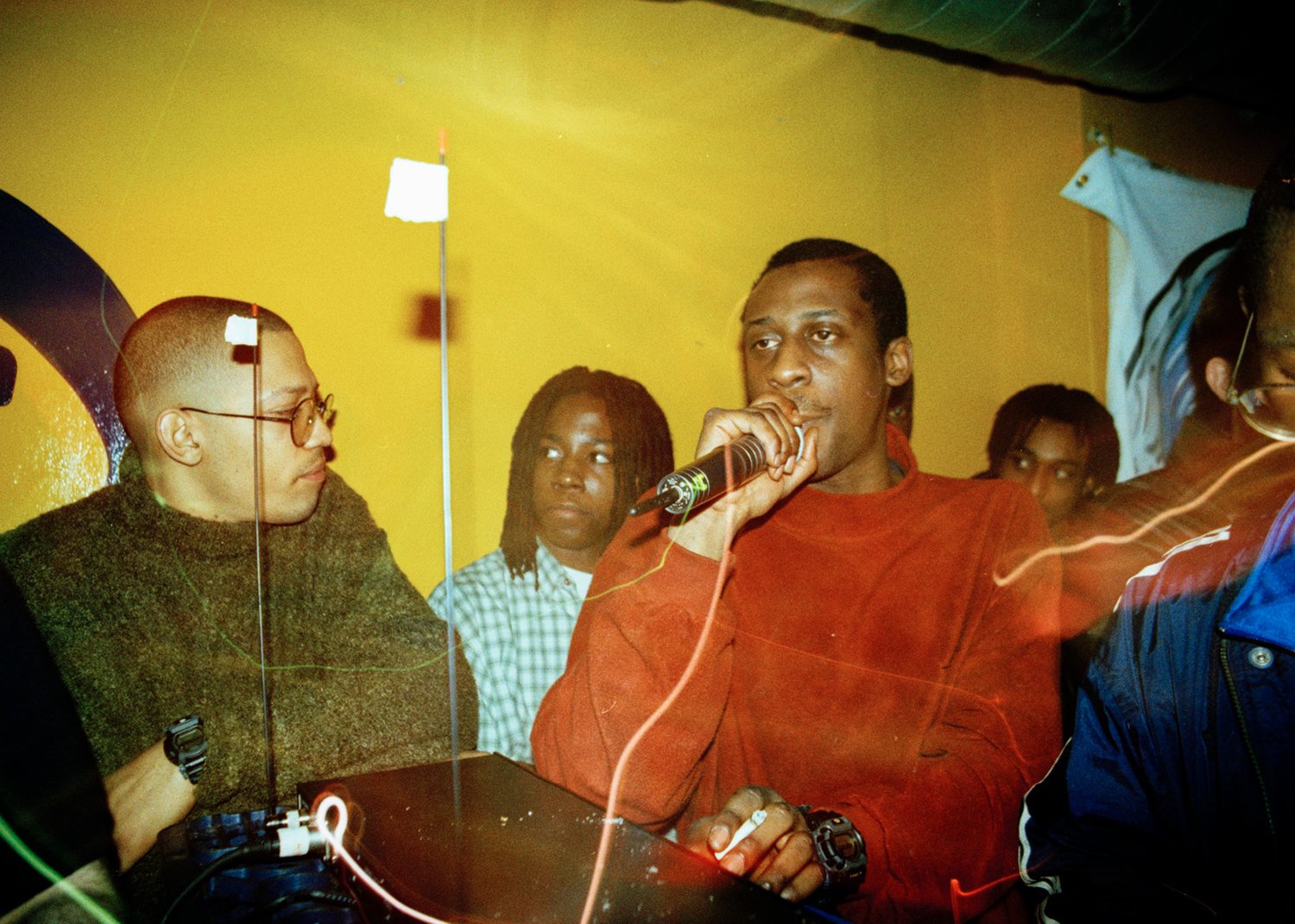 MC in red shirt holding a microphone in front of a yellow wall with other people around in a club.