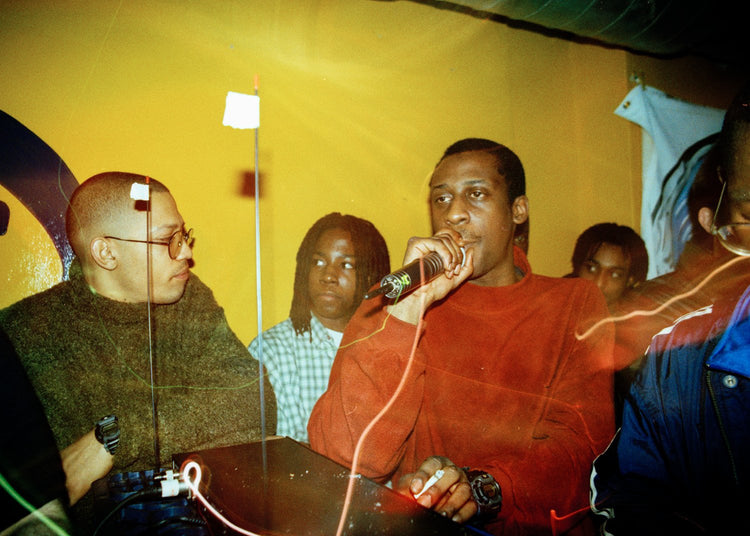 MC in red shirt holding a microphone in front of a yellow wall with other people around in a club.