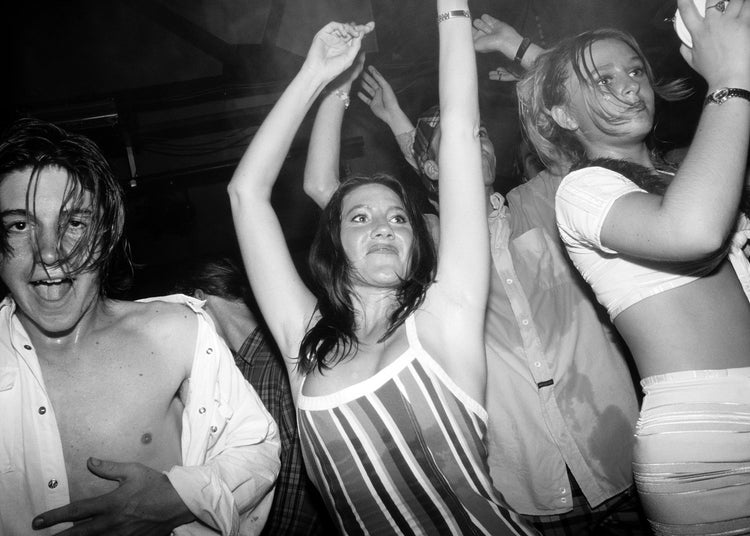 Black and white photograph of people dancing at a party with one person raising their arms.