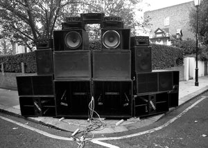 Stack of large speakers on a street corner with trees and buildings in the background, Notting Hill, London