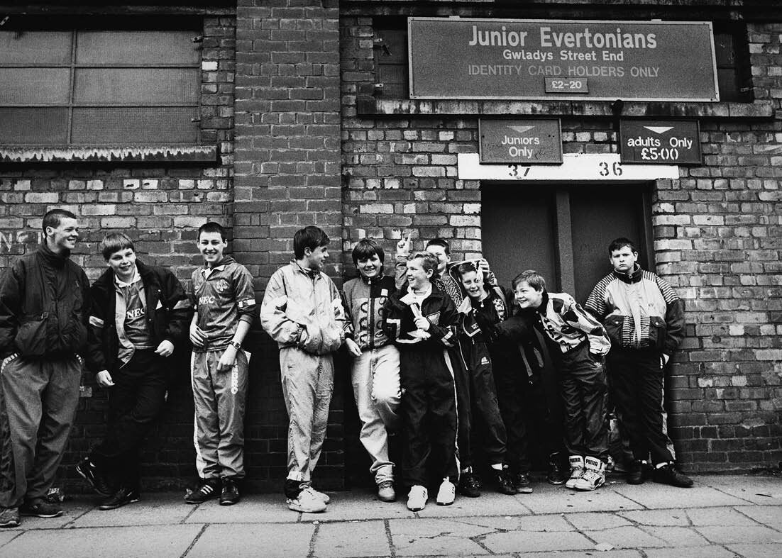 Group of young people posing in front of a brick building with a sign for 'Junior Evertonians'.