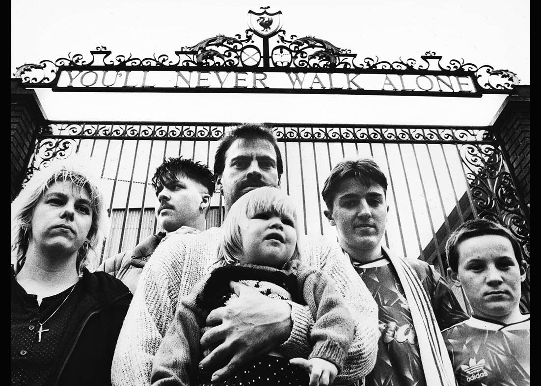 Group of people standing in front of Anfield gate with 'You'll Never Walk Alone' inscription