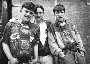 Three young men wearing Barcelona football club jerseys and scarves in front of a metal door.