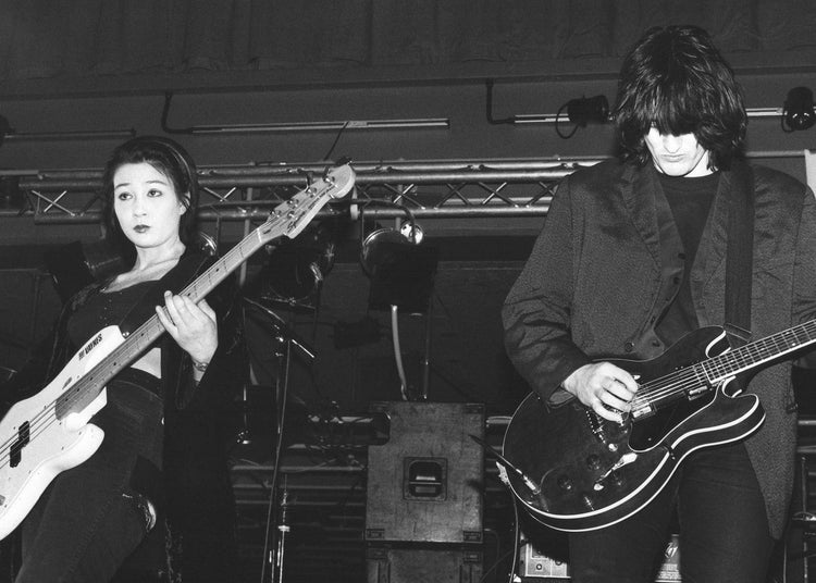 Two musicians playing electric guitars on a stage with musical equipment in the background.