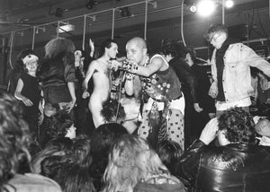 Black and white photo of a lively gig scene with people dancing on stage and interacting.