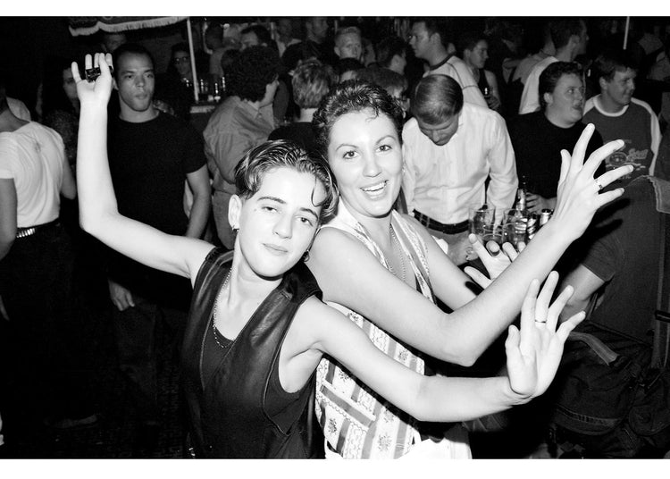 Two people posing together at a club with people in the background.