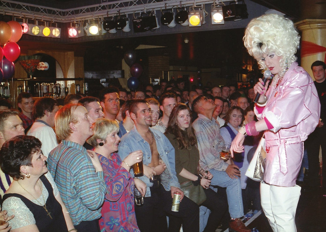 Drag queen in a pink outfit with a microphone addressing an audience in a club setting