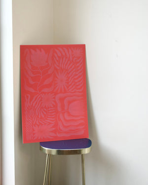 A rectangular red art print or panel with a tonal floral and leaf pattern, leaning against a white wall on a small stool with a blue seat and gold legs.