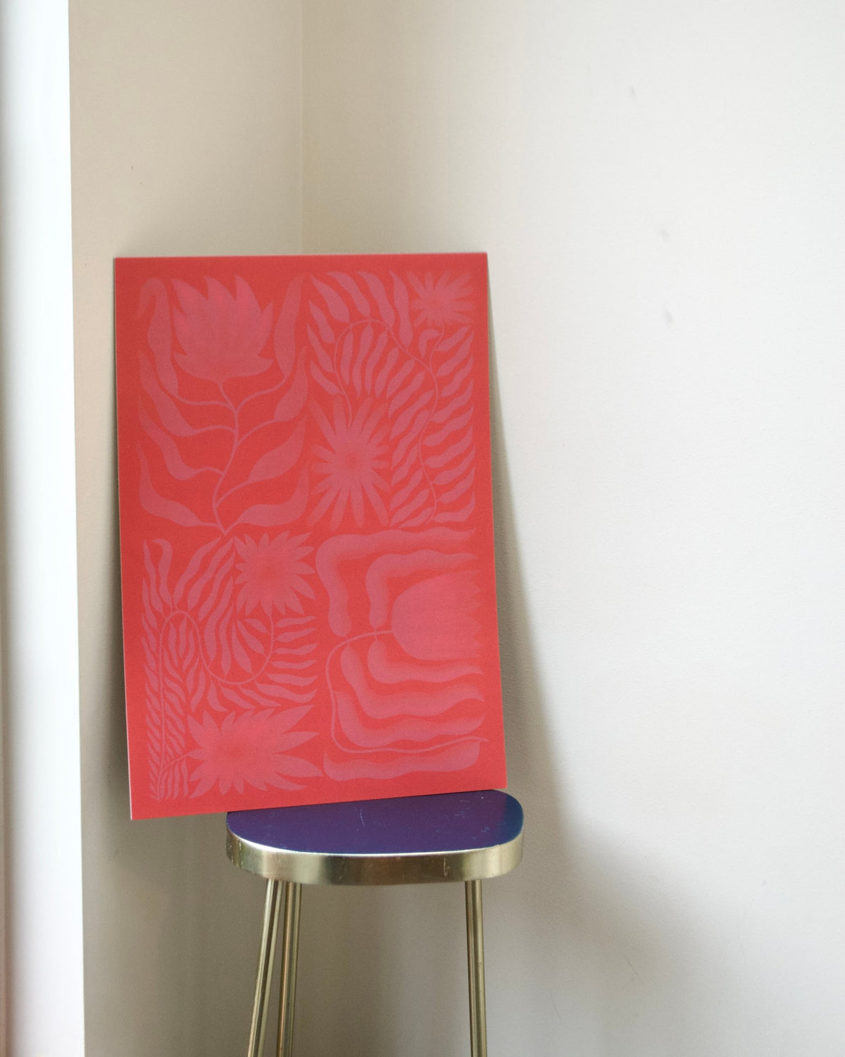 A rectangular red art print or panel with a tonal floral and leaf pattern, leaning against a white wall on a small stool with a blue seat and gold legs.