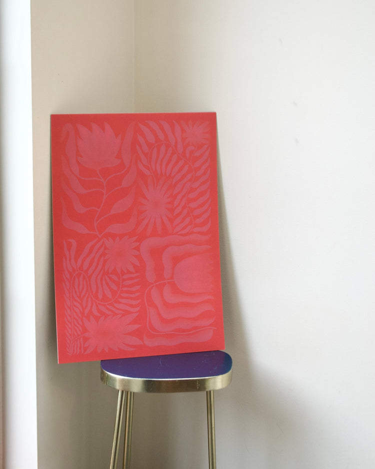A rectangular red art print or panel with a tonal floral and leaf pattern, leaning against a white wall on a small stool with a blue seat and gold legs.
