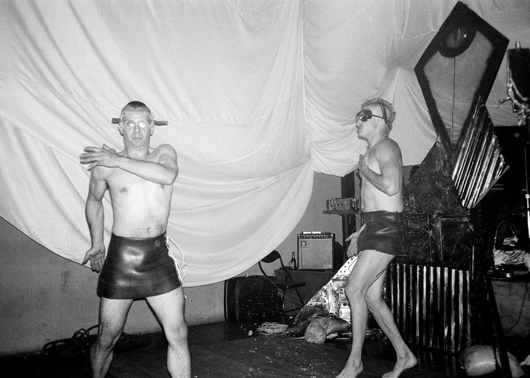 Two people in leather outfits dancing in a room with a curtain and equipment.