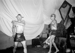 Two people in leather outfits dancing in a room with a curtain and equipment.