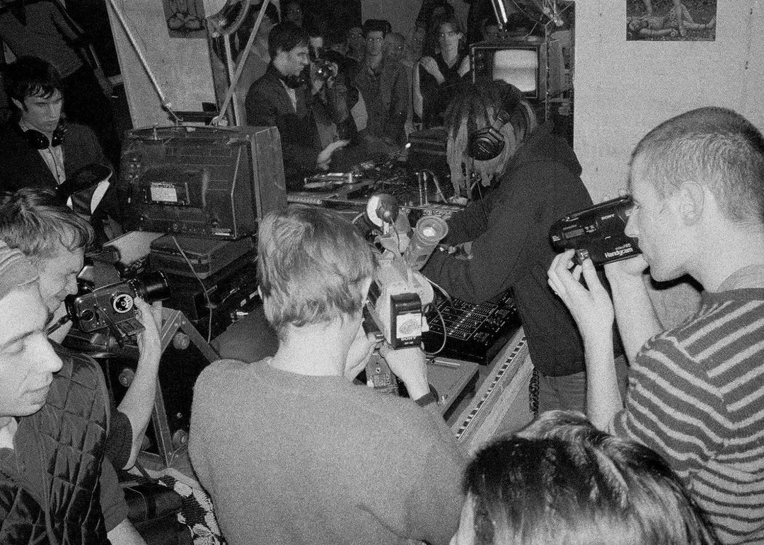 Black and white scene of people with electronic music and video cameras in a crowded room.