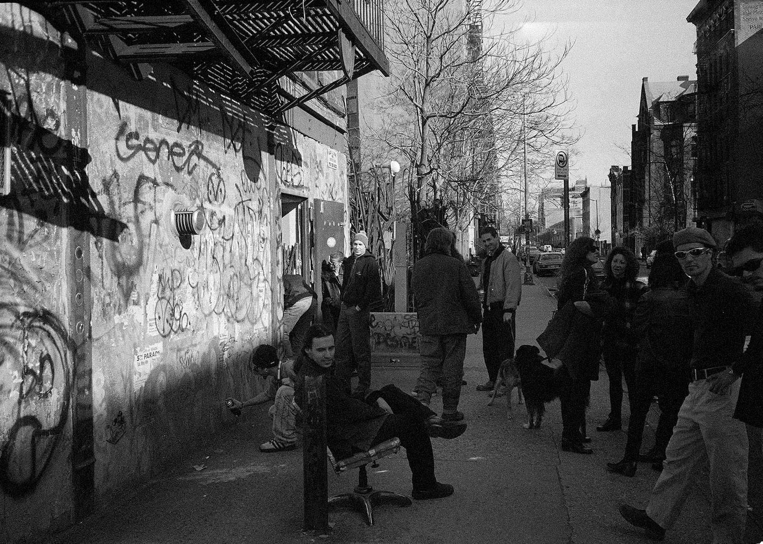 Black and white street scene with people on a New York sidewalk and graffiti on a wall.