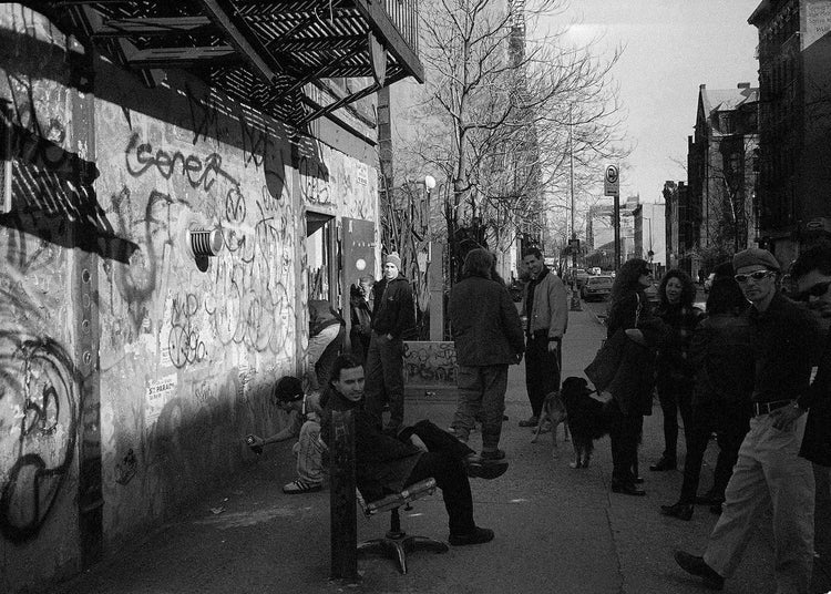 Black and white street scene with people on a New York sidewalk and graffiti on a wall.