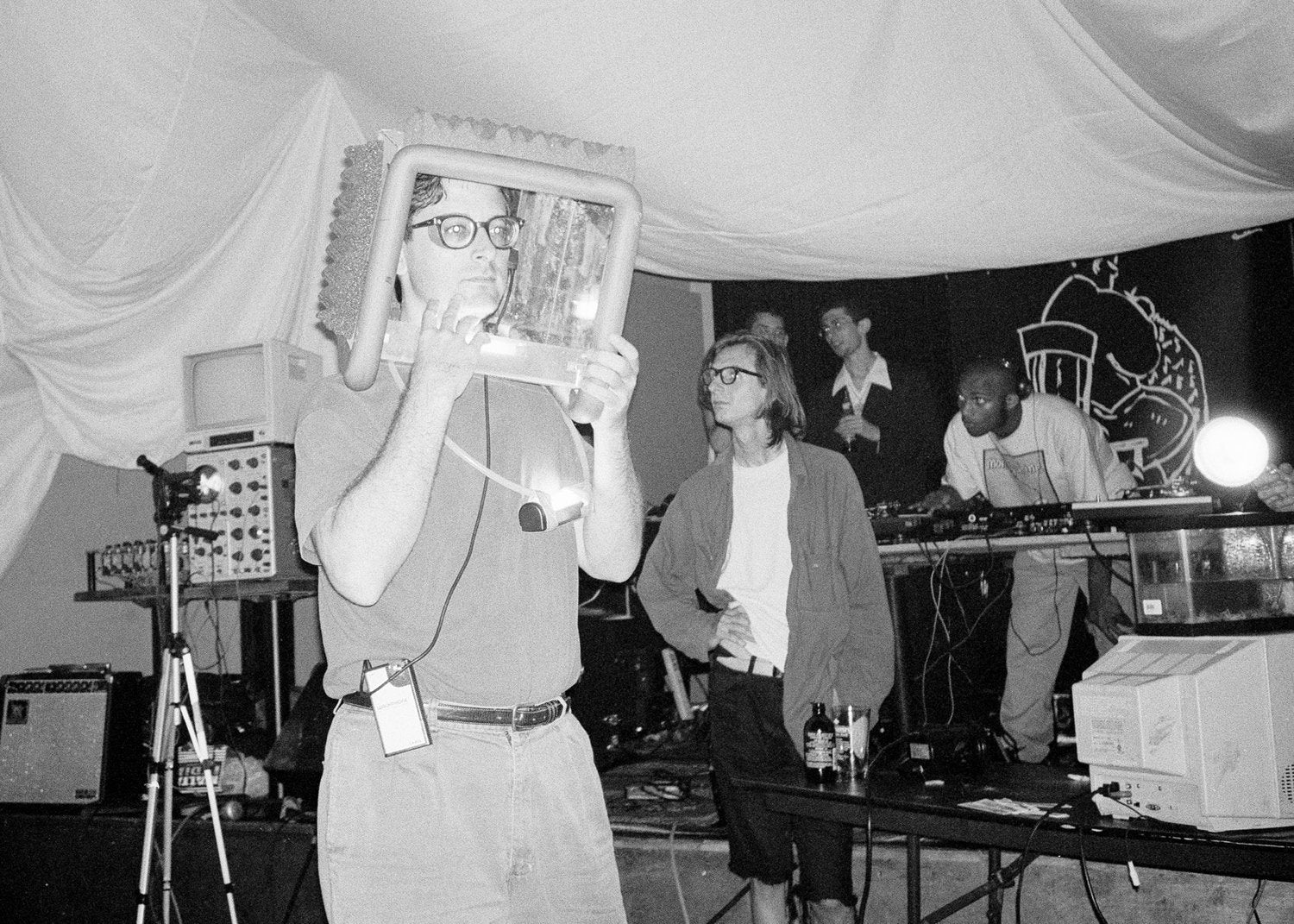 Person holding a framed object in front of their face at a music event with other people and equipment.