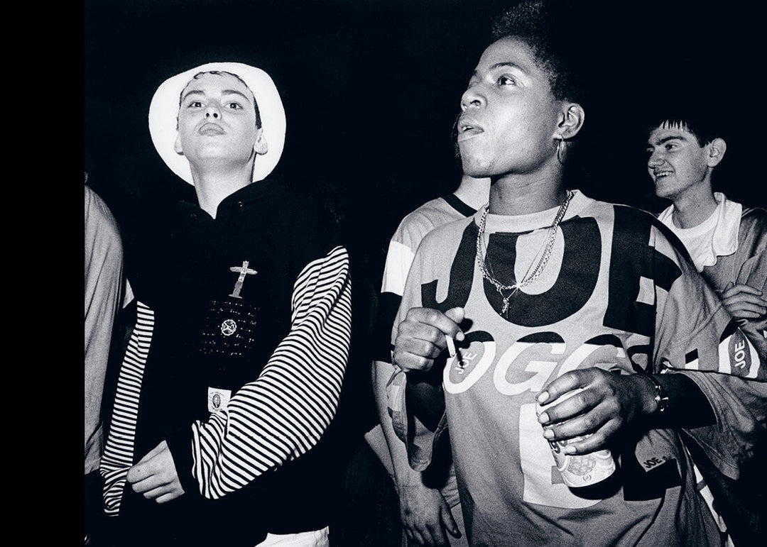 Black and white photo of a group of people, one holding a can wearing a chain, one wearing a white hat.