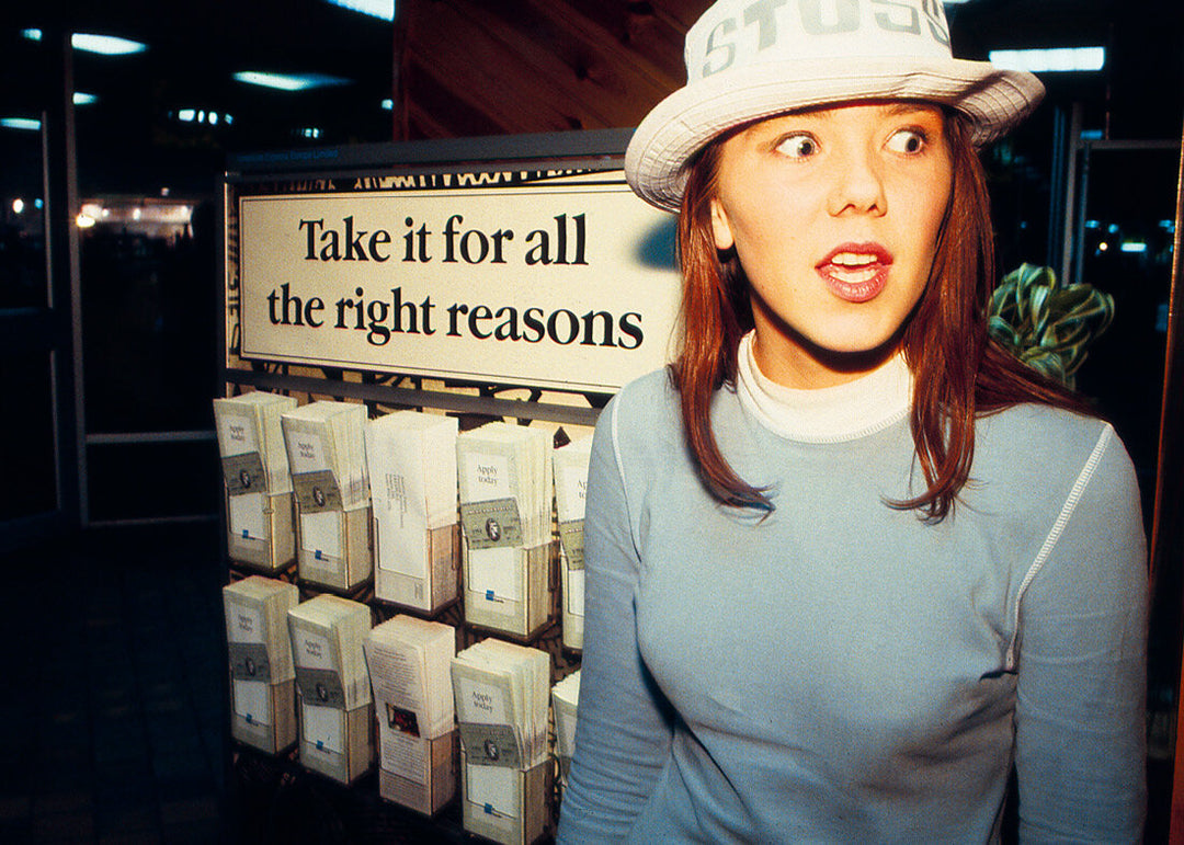 Woman wearing a white bucket hat with a surprised expression, standing in front of a display with boxes and a sign "Take it for all the right reasons"