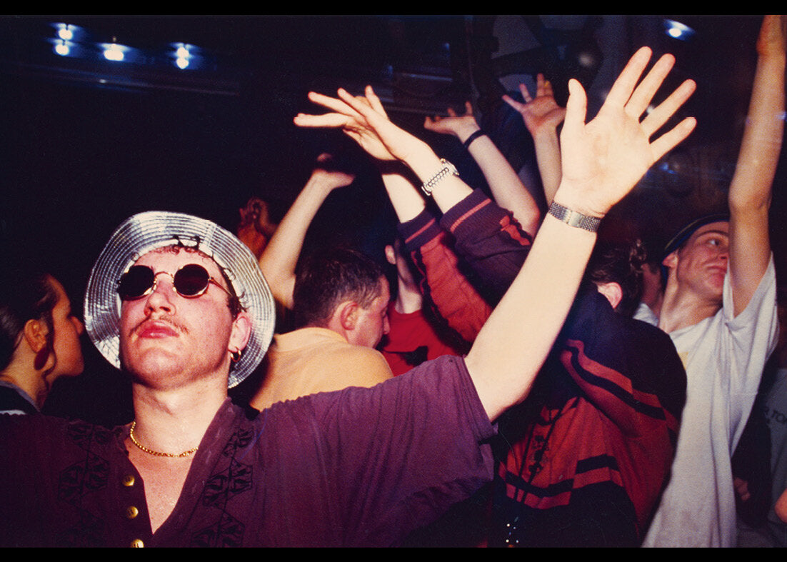 Person with raised arms at a rave, wearing sunglasses and a hat.