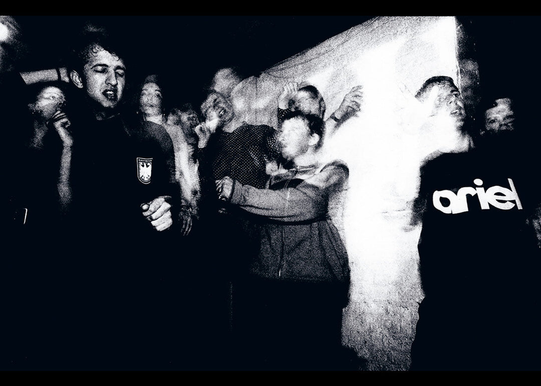 Black and white image of a group of people at rave with one person wearing a 'ariel' shirt.