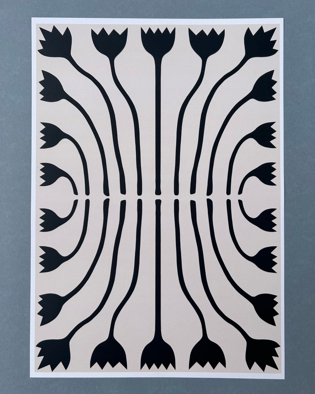 Tilly Worth Tulips A3 print featuring a symmetrical black floral pattern on a light beige background, bordered in white and photographed against a muted blue-grey backdrop.