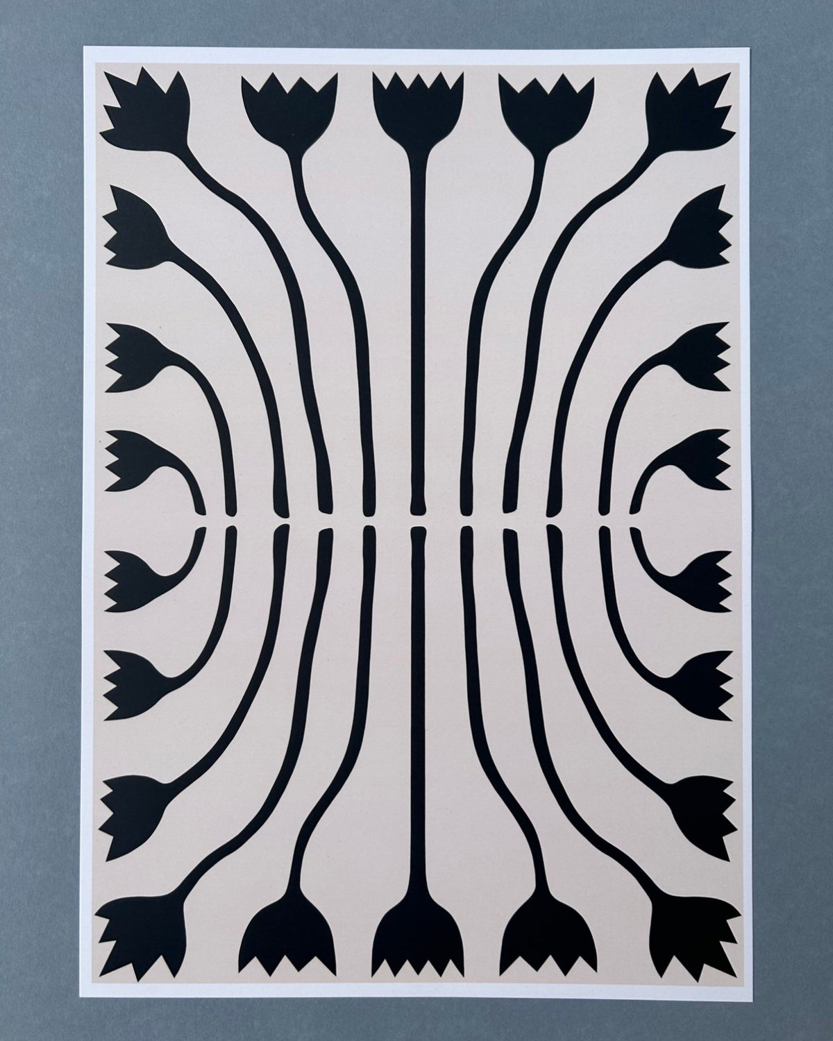 Tilly Worth Tulips A3 print featuring a symmetrical black floral pattern on a light beige background, bordered in white and photographed against a muted blue-grey backdrop.