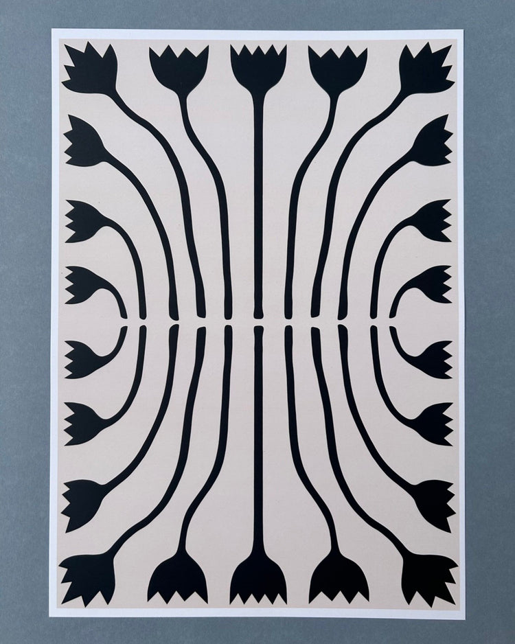 Tilly Worth Tulips A3 print featuring a symmetrical black floral pattern on a light beige background, bordered in white and photographed against a muted blue-grey backdrop.
