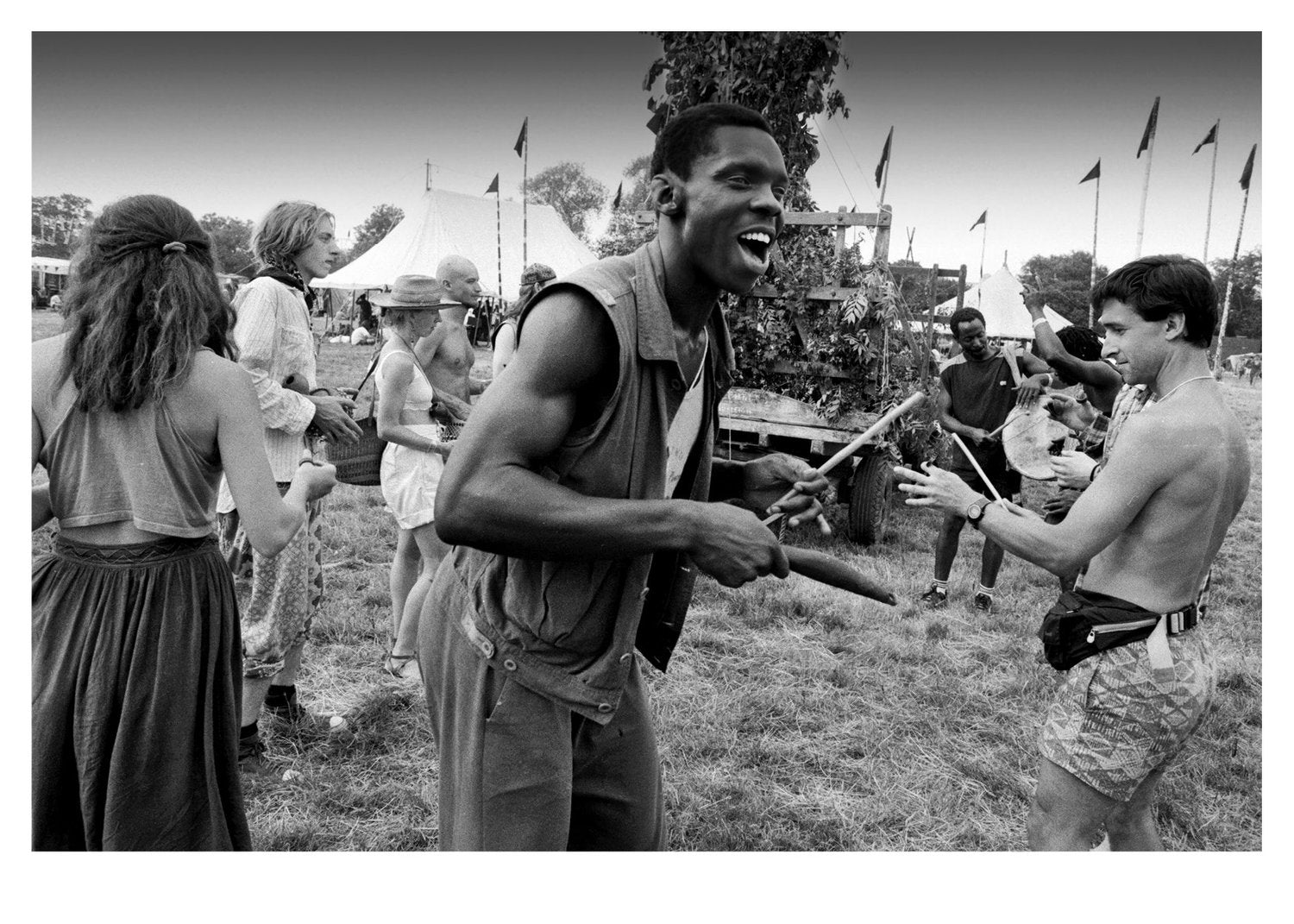 Black and white scene of a man in a sleeveless shirt holding a stick, interacting with another person in a grassy outdoor festival setting.